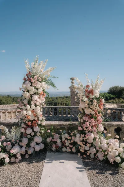 Ceremony setup featuring outdoor ceremony decor with a white aisle runner, framed by a stone balustrade on a terrace with trees and distant hills.