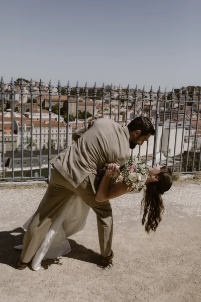 Wedding couple portrait of groom dipping bride for a kiss, her rose bouquet in hand, with city skyline beyond an iron railing overlook