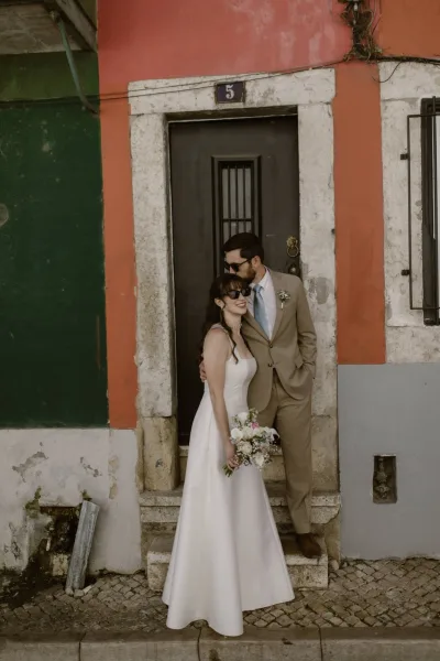 Couple portrait of bride and groom sunglasses as he kisses her forehead, bouquet in hand, by a colorful doorway on cobblestones