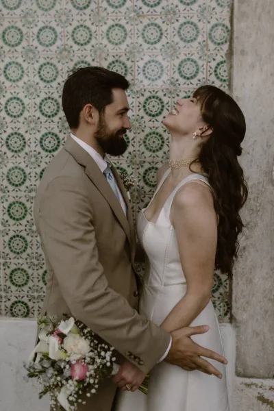 Couple portrait of bride and groom portrait laughing close together, bride in satin square-neck gown with bouquet, against patterned tile wall
