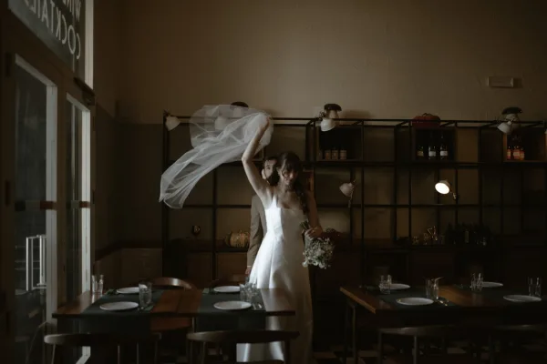Bride portrait with a long bridal veil and white bouquet, wearing a strapless gown in a softly lit restaurant with set tables