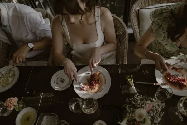 Wedding reception dinner overhead view of a bride eating at reception, white wedding dress beside plates, glassware, rings, bouquet on dark wood table