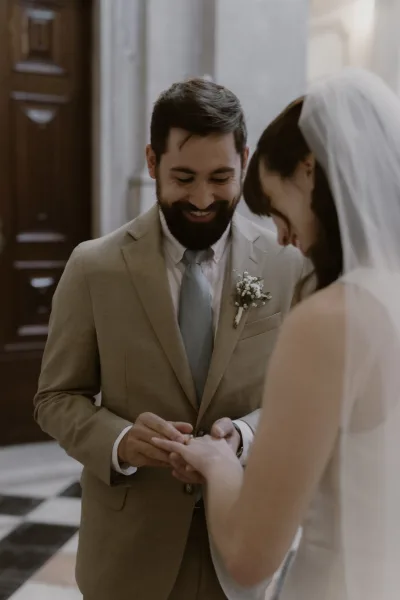 Ring exchange as groom places a wedding ring on the bride’s hand, veil and boutonniere visible in an indoor hallway with columns