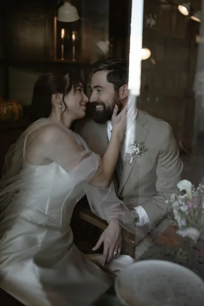 Couple portrait of bride and groom close up, bride touching groom’s bearded face in a moody restaurant with window reflection and pendant lights