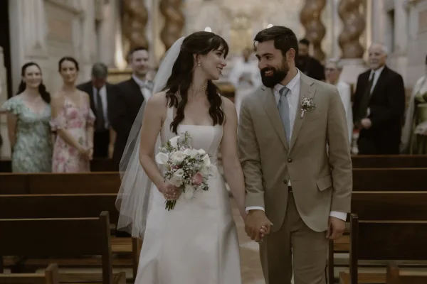 Recessional moment as bride and groom walk down aisle holding hands, bride with white bouquet and veil in church pews among guests