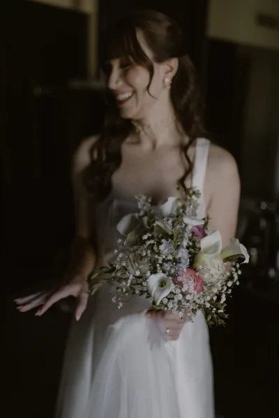 Bridal portrait of a laughing bride holding bouquet of calla lilies, baby's breath and pink roses in soft window light by a dark doorway