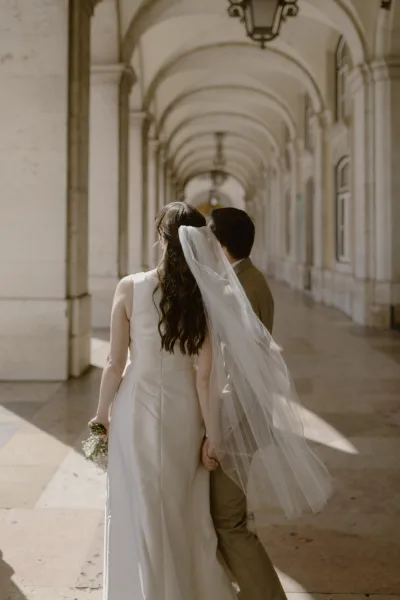 Couple portrait of bride and groom walking away, holding hands as her long veil trails through a sunlit stone colonnade corridor