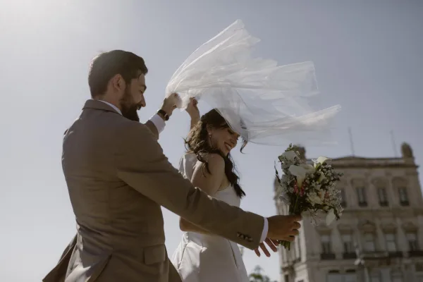 Couple portrait of bride and groom walking as he lifts her windy veil, bride holding bouquet in sunlight by a historic building