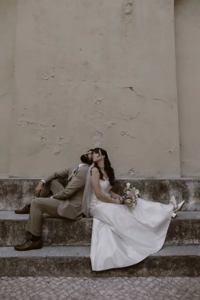 Couple portrait of bride and groom sitting back to back on stone steps, bride in veil holding a rose bouquet beside groom in tan suit