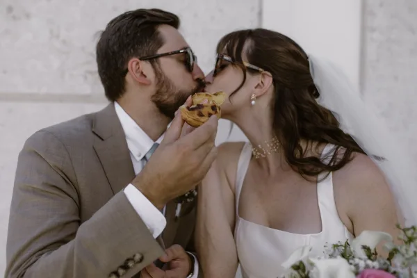 Wedding kiss as bride and groom kiss in sunglasses, her veil draped over a satin gown against a white stone wall backdrop