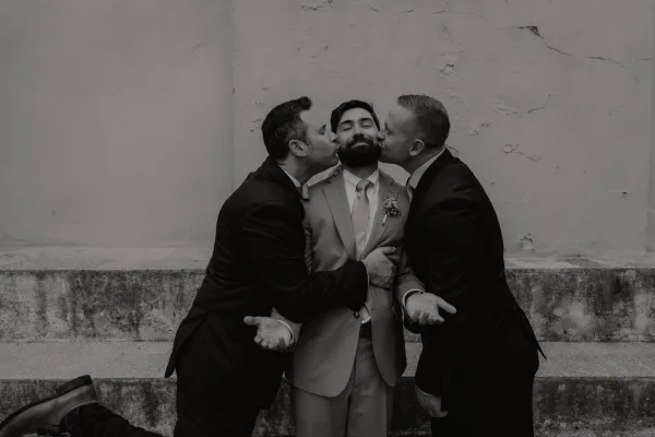 Groomsmen portrait of groom with groomsmen kissing his cheeks, wearing suits with boutonniere accents by a stucco wall and stone steps
