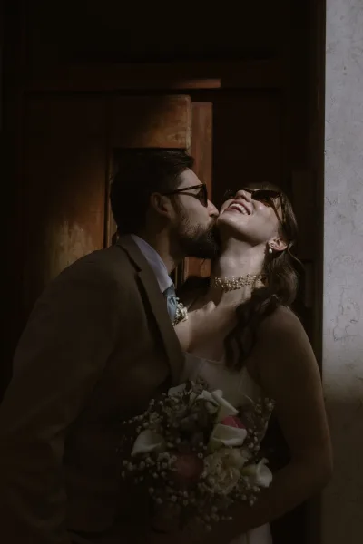 Wedding couple portrait of bride and groom kiss, both in sunglasses, with bride holding rose bouquet at rustic wooden doorway in warm light