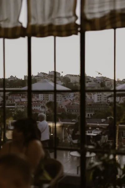 City skyline view from a rooftop terrace view through window panes, with café tables and railing overlooking hillside rooftops and trees