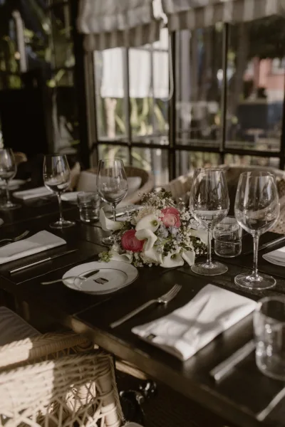 Reception tablescape with wedding table setting, low floral centerpiece, wine glasses, and white napkins by glass windows and greenery