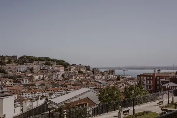 Cityscape view with rooftop city skyline beyond an iron fence, benches and street lamps overlooking terracotta rooftops, river and distant bridge