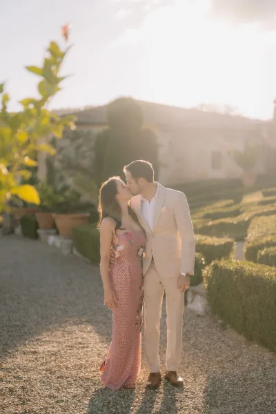 Couple kiss portrait at golden hour, her strapless embellished gown catching light on a gravel path by a stone villa framed with formal hedges.