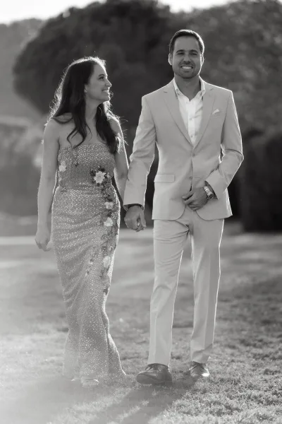 Couple portrait of bride and groom walking hand in hand across a garden lawn, her strapless gown adorned with floral appliqués.
