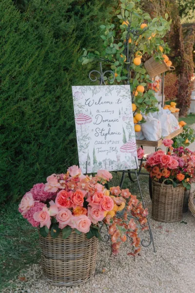 Wedding welcome sign styled as outdoor wedding signage, trimmed with citrus branches beside garden hedges and a gravel path.