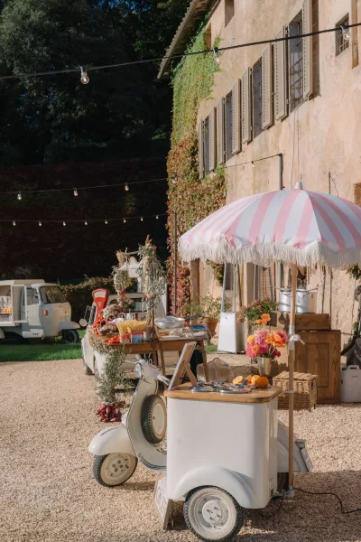 Wedding gelato cart styled as a dessert station at a gravel courtyard, shaded by a striped umbrella beside an ivy-clad stone facade.