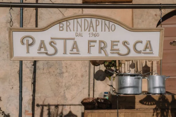 Pasta bar sign marking a wedding pasta station, hung on a rod with hanging utensils against a stucco wall near a soft-shadowed doorway arch.