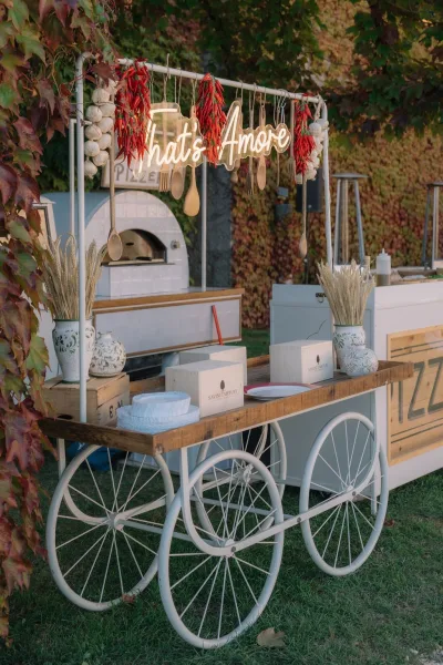 Wedding pizza station with mobile pizza oven catering, set beneath a glowing neon sign by an ivy-covered wall on a green lawn.