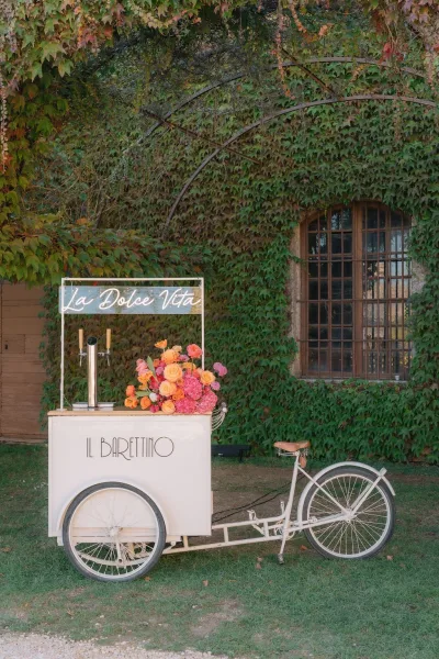 Wedding mobile bar styled as a bicycle bar cart, glowing with a neon sign against an ivy wall and arched window on a grass lawn