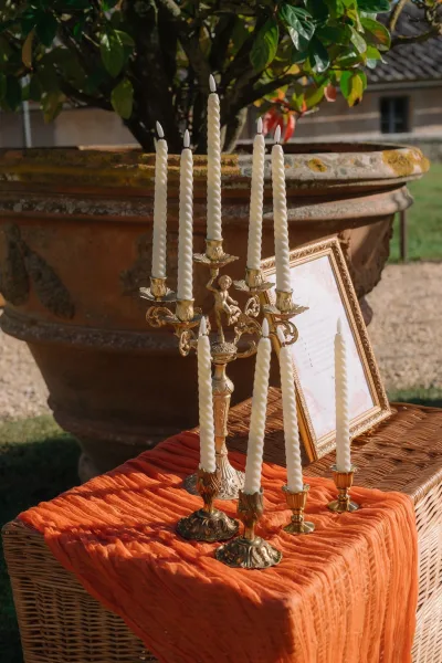 Wedding candle display featuring a gold candelabra centerpiece on an orange fabric runner, set by a terracotta planter and green foliage.
