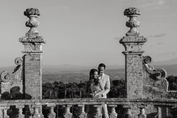 Couple portrait with the groom hugging the bride, her bridal gown against a stone balustrade and ornate pillars, hills fading under open sky.