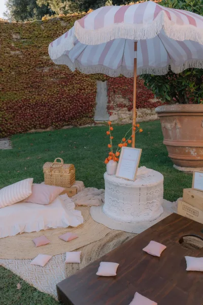 Outdoor lounge setup for a wedding lounge area anchored by a striped fringe umbrella, set on a grass lawn beside an ivy-covered wall.