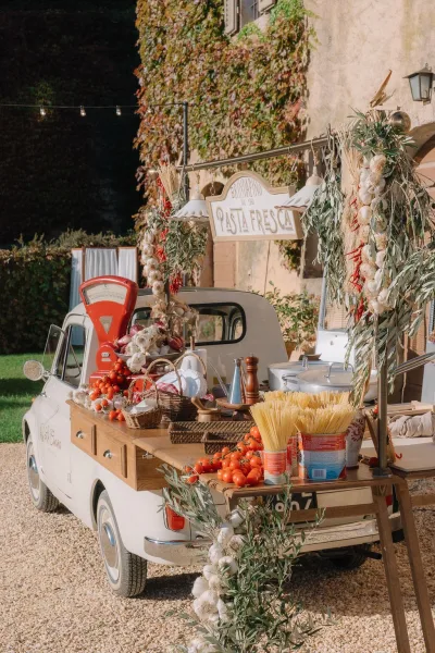 Pasta bar setup at a vintage car food bar, trimmed with garlic garlands, against an ivy-covered wall in a gravel courtyard.