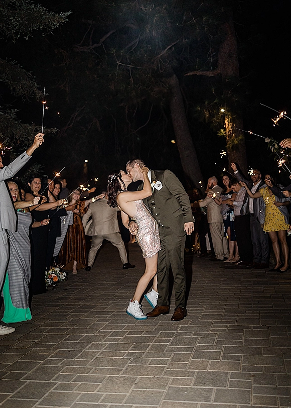 Wedding send off with a sparkler send off tunnel as the bride in a mini dress and sneakers kisses her groom under string lights at night