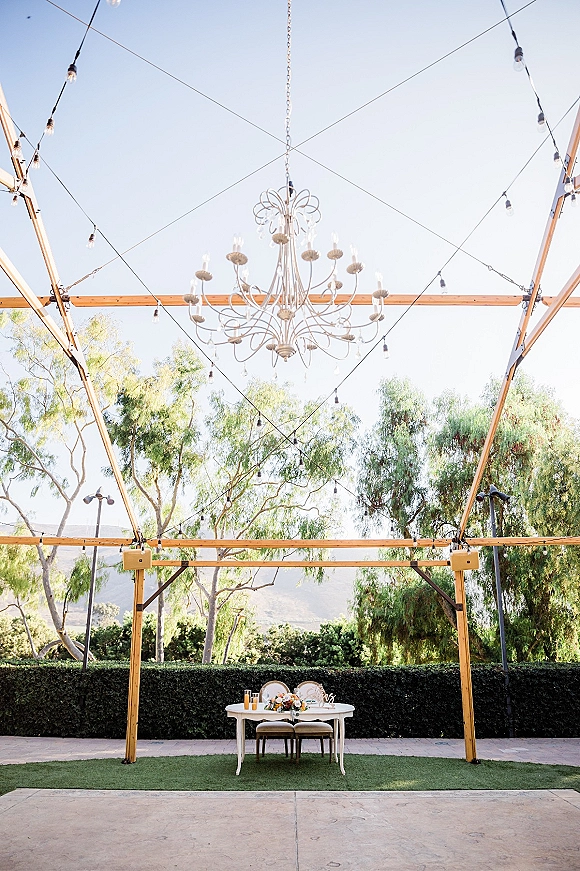 Sweetheart table decor with floral centerpiece and candles beneath a white chandelier and string lights on an outdoor patio lawn setting