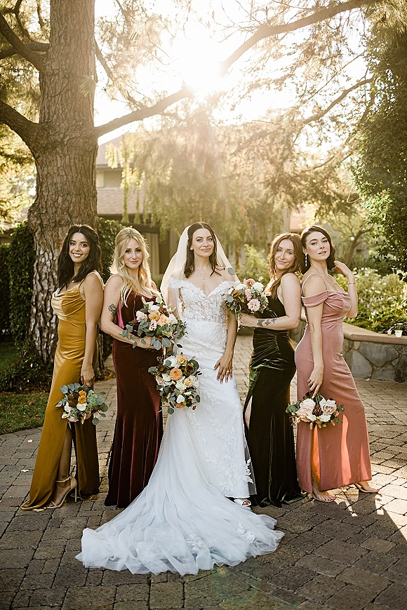 Bride with bridesmaids in a bridesmaid group photo holding bouquets, bride in veil and wedding dress, standing in a sunny garden courtyard