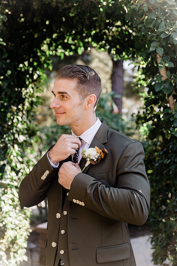 Groom portrait of a man adjusting his tie in an olive green three-piece suit with boutonniere beneath a greenery arch in dappled sunlight