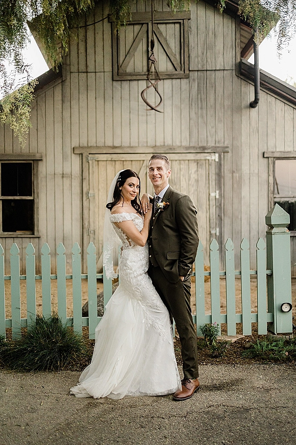 Couple portrait of bride in off-the-shoulder lace gown and veil holding groom’s lapel beside barn doors and a white picket fence