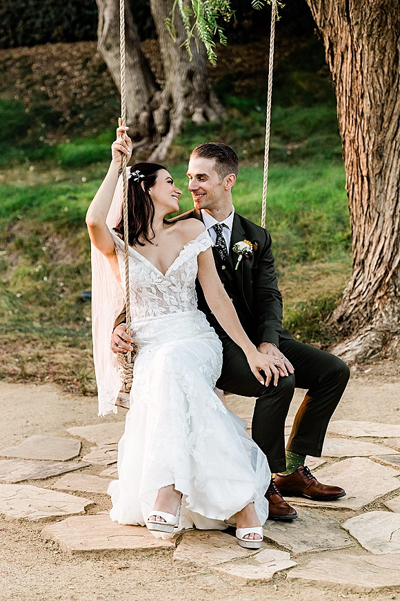 Couple portrait of bride and groom on swing, holding hands as her long veil drapes over a strapless lace dress on a grassy lawn