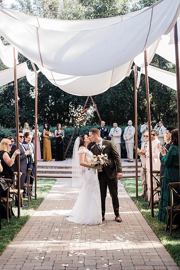 Wedding kiss as bride and groom embrace under a white fabric canopy, bride in lace dress with veil holding bouquet on brick aisle outdoors