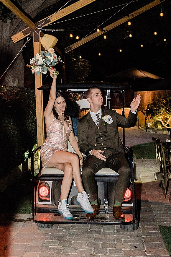 Wedding exit as bride in sequin mini dress holds bouquet beside groom on a golf cart under string lights at night pergola walkway