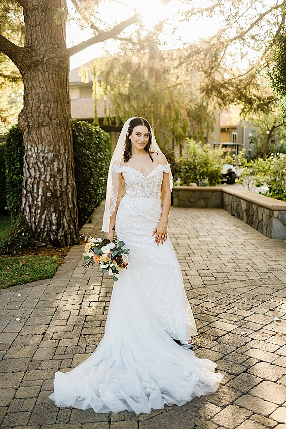 Bridal portrait of a bride in an off-shoulder lace wedding dress holding a greenery bouquet, cathedral veil flowing on a sunlit garden walkway