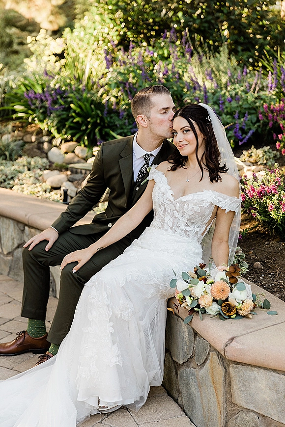 Couple portrait of bride and groom sitting on a stone bench as he kisses her forehead, with veil and bouquet in a garden setting