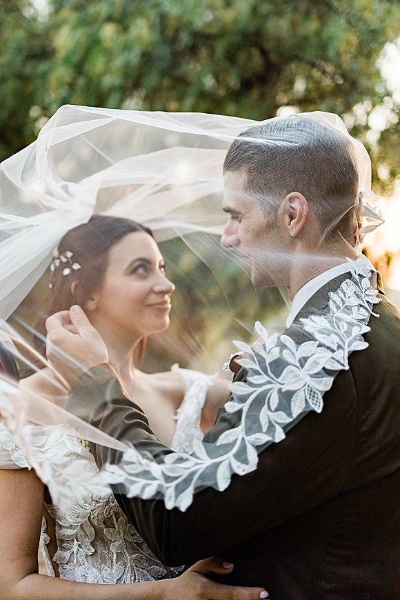 Couple portrait with a wedding veil draped over them as the groom holds the bride’s face, sunlight filtering through trees behind