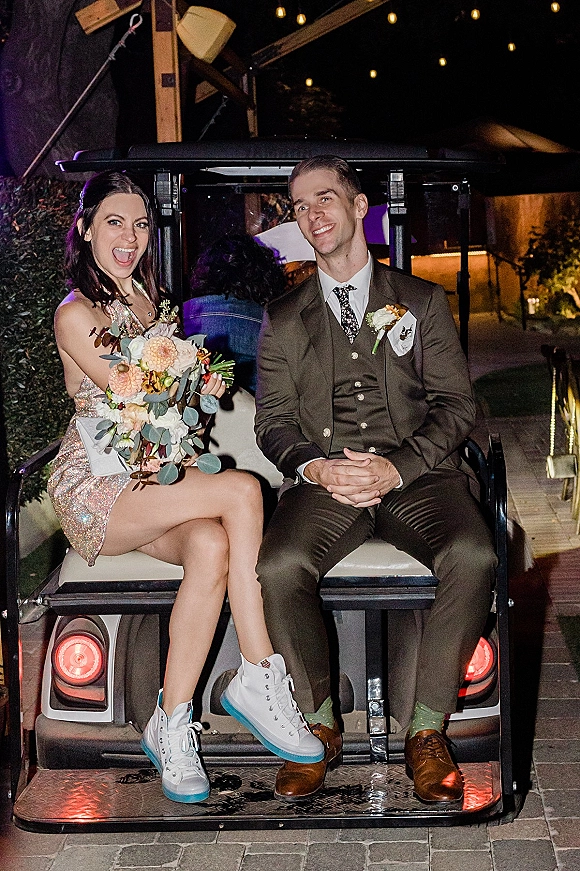 Couple portrait of newlyweds on a wedding golf cart exit under string lights at night, bride in sequin dress holding bouquet beside groom in suit