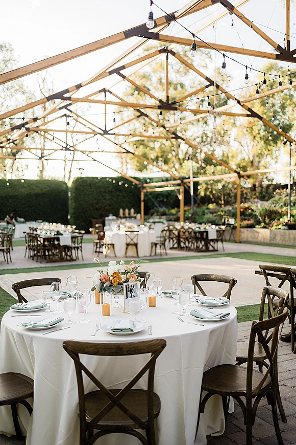 Reception tablescape at an outdoor wedding reception with a round white tablecloth table, floral centerpiece, votive candles, and string lights overhead