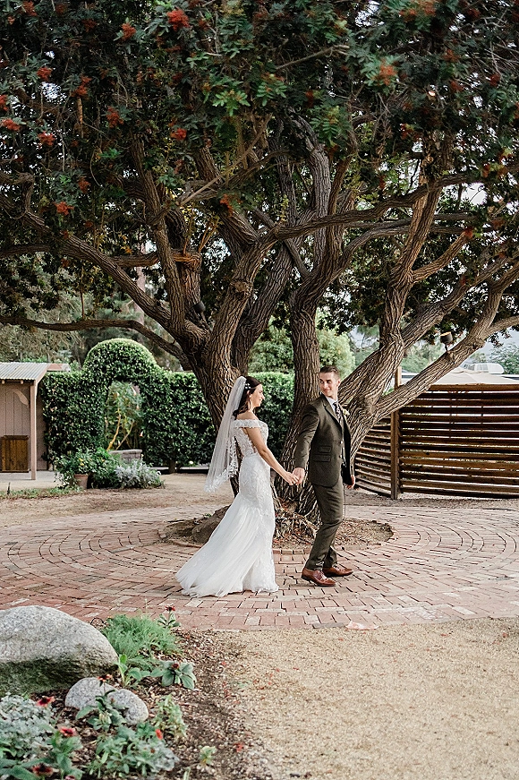 Couple portrait of bride and groom holding hands, her lace gown and veil trailing as they walk under a large tree in a brick courtyard