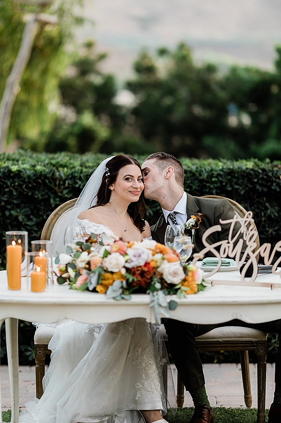 Wedding sweetheart table with bride in veil and groom in suit kissing beside candles and low floral centerpiece on stone patio by garden hedge