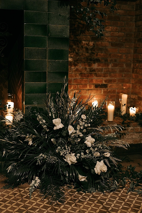 Wedding ceremony florals with an altar floral arrangement of white roses, greenery and palm fronds, surrounded by pillar candles by a brick fireplace