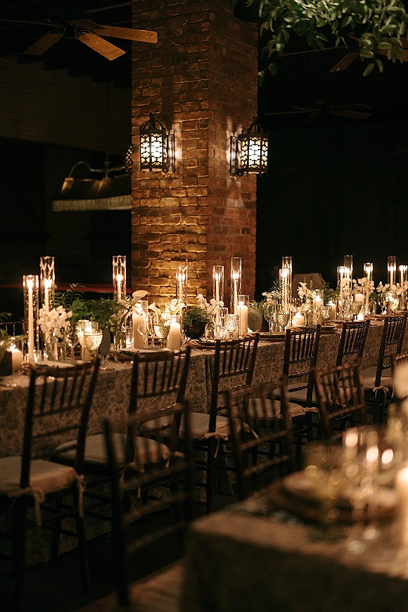 Reception tablescape with candlelit wedding reception glow, taper candles and white florals on a patterned tablecloth in a moody brick room