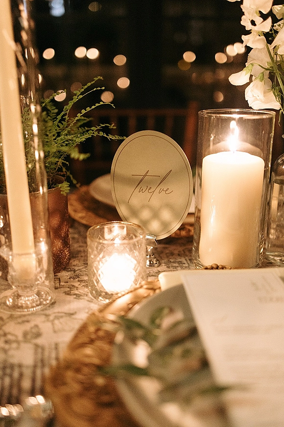Reception tablescape with a candlelit wedding table, pillar candles in hurricanes, greenery and white flowers, gold chargers, and string lights at night