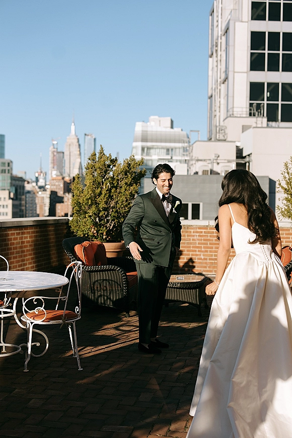 First look moment as bride in a white satin wedding dress approaches groom in black tuxedo on a rooftop terrace with city skyline backdrop