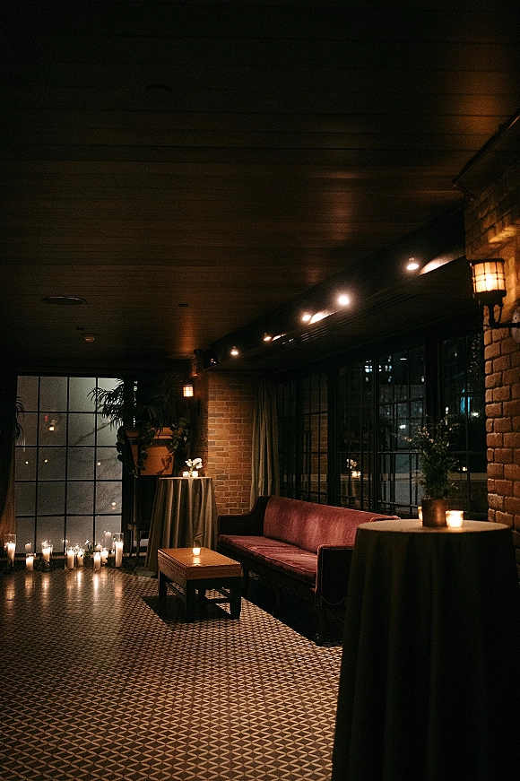 Wedding lounge area with reception lounge seating, burgundy velvet sofa and candlelit cocktail tables against brick walls and large windows at night
